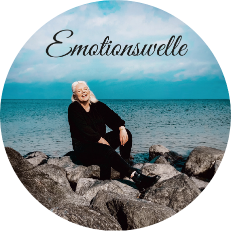 Frau sitzt auf Steinen am Meer und sinkt. Im Himmel ist ein Schriftzug mit dem Text Emotionswelle